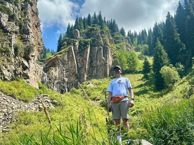 A man wearing a fanny pack standing in a green mountain valley with a waterfall.