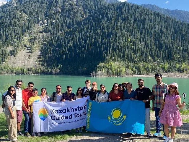 Un grupo de turistas sosteniendo la bandera de Kazajistán posando junto a un lago con montañas al fondo.